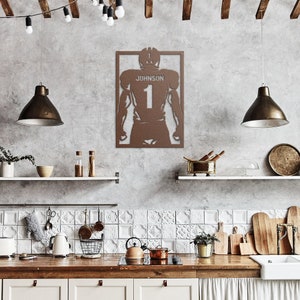 Pu&ograve; includere: Una silhouette di un giocatore di football americano in metallo marrone con il nome "Johnson" e il numero "1" sul retro. La silhouette &egrave; montata su una parete bianca in un ambiente da cucina.