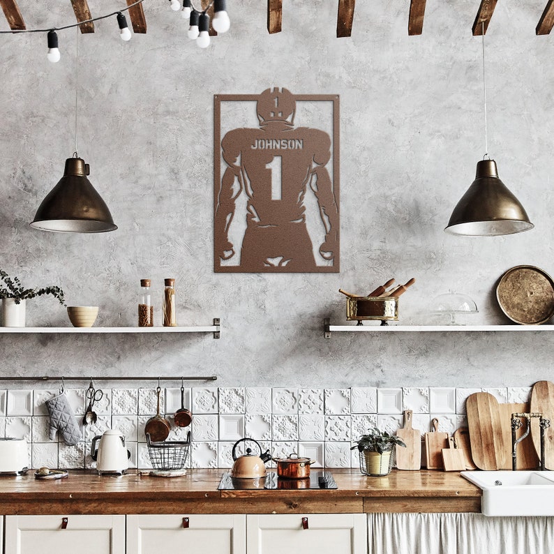 Pu&ograve; includere: Una silhouette di un giocatore di football americano in metallo marrone con il nome "Johnson" e il numero "1" sul retro. La silhouette &egrave; montata su una parete bianca in un ambiente da cucina.