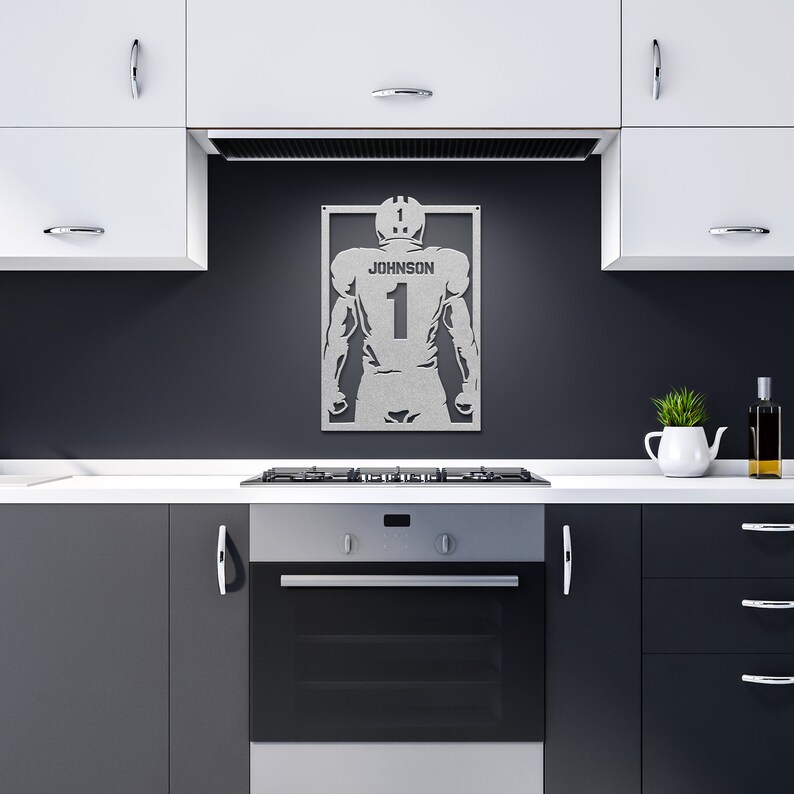Pu&ograve; includere: Una silhouette in metallo di un giocatore di football americano con il nome "JOHNSON" e il numero "1" sul retro. La silhouette &egrave; montata su una parete grigio scuro in un ambiente da cucina.