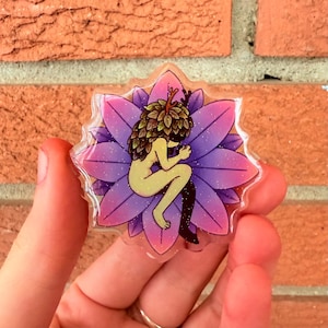 May include: A clear, star-shaped sticker featuring a seated figure with a leafy head and antlers, positioned within a purple and pink flower. The figure is nude with a black leg. The background is a brick wall.