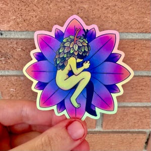 May include: A holographic sticker featuring a seated figure with a leafy head and small antlers, cradled within a vibrant, multi-colored flower. The figure has a pale yellow skin tone, and is wearing a black sock on one leg. The flower petals transition from purple to pink.
