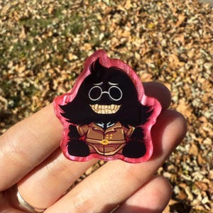 May include: A red-edged, star-shaped pin featuring a cartoon character with black hair, round glasses, and a wide, toothy grin. The character wears a brown jacket with gold buttons. The pin is held against a background of fallen leaves.