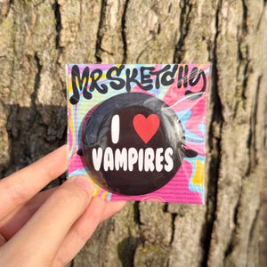May include: A black button with the text "I ❤️ VAMPIRES" and a red heart, packaged on a colorful card. The button is labeled "Mr Sketchy" in a playful font. The background is a tree trunk.
