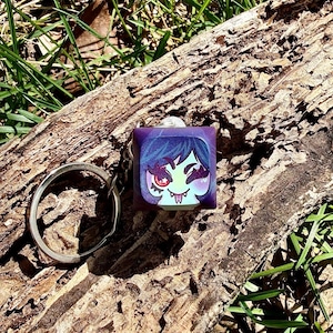 May include: A purple square keychain with a cartoon illustration of a character with blue hair, a red eye, and fangs. The keychain is attached to a silver metal ring. The background is a wooden log with green grass.