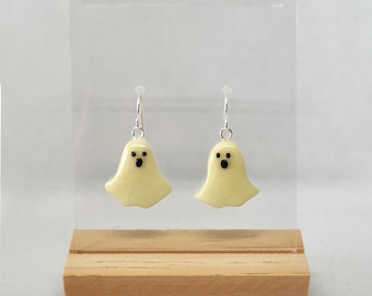 Op de afbeelding: Een paar spookvormige oorbellen met zwarte ogen. De oorbellen zijn gemaakt van wit hars en hangen aan zilveren haakjes. De oorbellen worden gepresenteerd op een houten standaard.