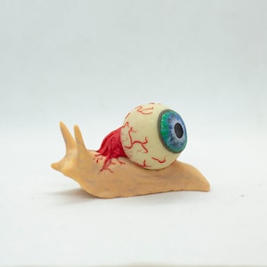 May include: A small, beige snail with a large, white eyeball on its back. The eyeball has red veins and a blue iris.