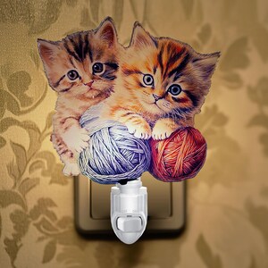 May include: A night light featuring two kittens playing with balls of yarn. The kittens are brown and white with blue eyes. The night light is white with an on/off switch.