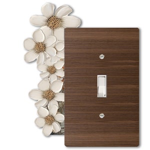 Puede incluir: Una tapa de interruptor de luz con un diseño de veta de madera y un interruptor de palanca blanco. La tapa está decorada con un diseño floral con flores blancas y centros marrones.