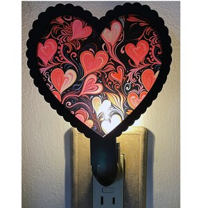 May include: A heart-shaped night light with a black scalloped border. The heart design features red, pink, and orange hearts on a black background with swirling patterns. The night light is plugged into a wall outlet.
