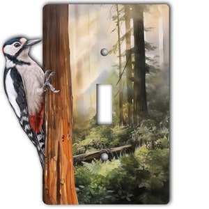 May include: A light switch cover featuring a watercolor painting of a woodpecker perched on a tree trunk in a misty forest. The background shows tall trees and lush greenery.