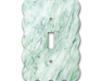 Op de afbeelding: Een decoratieve lichtschakelaar cover met een golvende rand. De cover heeft een gemarmerd patroon in tinten wit en groen. Het heeft een enkele schakelaar opening en twee schroefgaten.