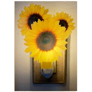 May include: A decorative night light featuring three bright yellow sunflowers with dark brown centers. The night light plugs into a wall outlet and has a yellow light cover. The sunflowers are arranged in a cluster, creating a cheerful and inviting ambiance.