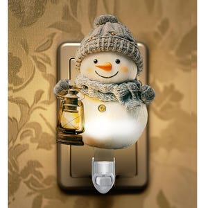 May include: A snowman night light with a gray knit hat and scarf, holding a lantern. The snowman has a carrot nose and a smiling face. The night light plugs into a wall outlet and has an on/off switch.