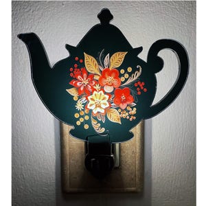May include: A teapot-shaped night light featuring a dark green background with a vibrant floral design in shades of red, orange, yellow, and gold. The central illustration displays detailed petals, leaves, and small berries in a symmetrical arrangement.