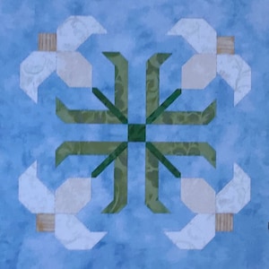 Pode incluir: Um bloco de colcha azul e branco com um desenho geométrico verde no centro. O desenho é rodeado por quatro formas de flores brancas com centros marrons.