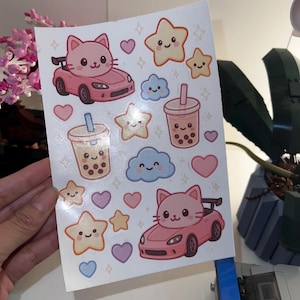 May include: A sticker sheet with various cute illustrations. Designs include pink cat cars, smiling stars, bubble tea, hearts, and fluffy clouds. The stickers have a kawaii aesthetic with a pastel colour palette.