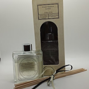 May include: A clear glass bottle with a black and white label that reads "Spa Relaxation Simple Elegance Reed Diffuser 4 oz.". The bottle is sitting on a white surface with a box of candles and a set of wooden diffuser sticks tied with a black and white ribbon.