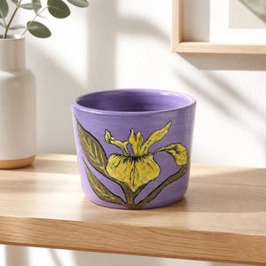 May include: A cylindrical ceramic planter with a smooth lavender finish, featuring a hand-painted illustration of a yellow iris flower with green leaves on the front. The pot has a wide, open top and a flat base, showcasing a vibrant floral design.