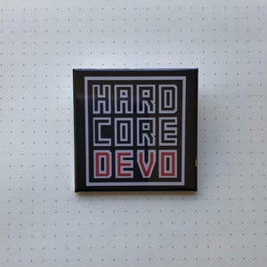 Könnte beinhalten: Ein quadratischer Knopf in Schwarzweiß mit dem Text "HARDCORE DEVO" in roten und schwarzen Buchstaben.
