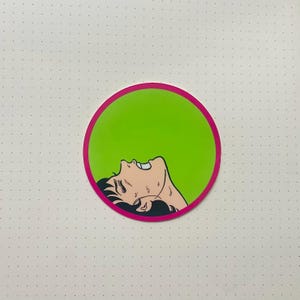 Puede incluir: Una pegatina circular verde con borde rosa que presenta una ilustración en blanco y negro de una persona con la cabeza echada hacia atrás y la boca abierta. La persona parece estar en apuros.