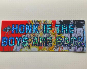 Op de afbeelding: Een sticker met een kleurrijke, stripboekachtige achtergrond met een blauwe en zwarte tekstoverlay die "HONK IF THE BOYS ARE BACK" zegt.
