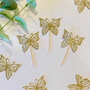 Pu&ograve; includere: Topper per cupcake a farfalla con glitter dorati. Questi topper decorativi presentano intricati disegni ritagliati e sono attaccati a bastoncini di legno. Le farfalle sono sparse su una superficie bianca, con foglie verdi sullo sfondo. Perfetti per una festa.