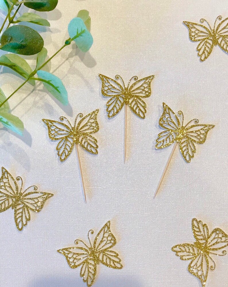 Pu&ograve; includere: Topper per cupcake a farfalla con glitter dorati. Questi topper decorativi presentano intricati disegni ritagliati e sono attaccati a bastoncini di legno. Le farfalle sono sparse su una superficie bianca, con foglie verdi sullo sfondo. Perfetti per una festa.
