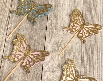 May include: Four butterfly-shaped cupcake toppers. Each butterfly has a glittery gold outline and is mounted on a wooden pick. The butterflies are in shades of blue, pink, and white.