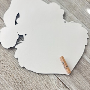 Pode incluir: Uma placa de madeira branca em forma de cora&ccedil;&atilde;o com uma borda decorativa e um pequeno prendedor de roupa de madeira preso. A placa tem um design recortado e um pequeno orif&iacute;cio para pendurar. O fundo &eacute; uma superf&iacute;cie de madeira.