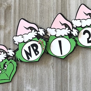 Pode incluir: Uma faixa festiva com tema Grinch. Cada recorte tem um rosto verde, um chap&eacute;u rosa com acabamento branco e um contorno preto. A faixa inclui as letras "NB", o n&uacute;mero "1" e o n&uacute;mero "2". Uma bengala de doces tamb&eacute;m &eacute; vis&iacute;vel.