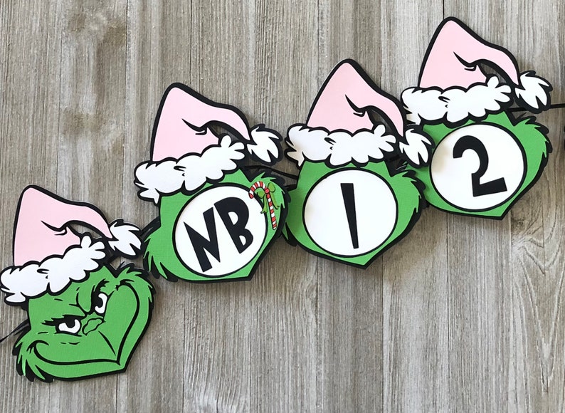 Pode incluir: Uma faixa festiva com tema Grinch. Cada recorte tem um rosto verde, um chap&eacute;u rosa com acabamento branco e um contorno preto. A faixa inclui as letras "NB", o n&uacute;mero "1" e o n&uacute;mero "2". Uma bengala de doces tamb&eacute;m &eacute; vis&iacute;vel.