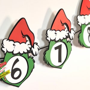 Pode incluir: Uma faixa festiva com recortes de rostos verdes com tema Grinch, com chap&eacute;us de Papai Noel vermelhos e acabamentos brancos. Cada recorte exibe um grande n&uacute;mero preto. A faixa inclui pequenos sinais com texto como "Whoville".