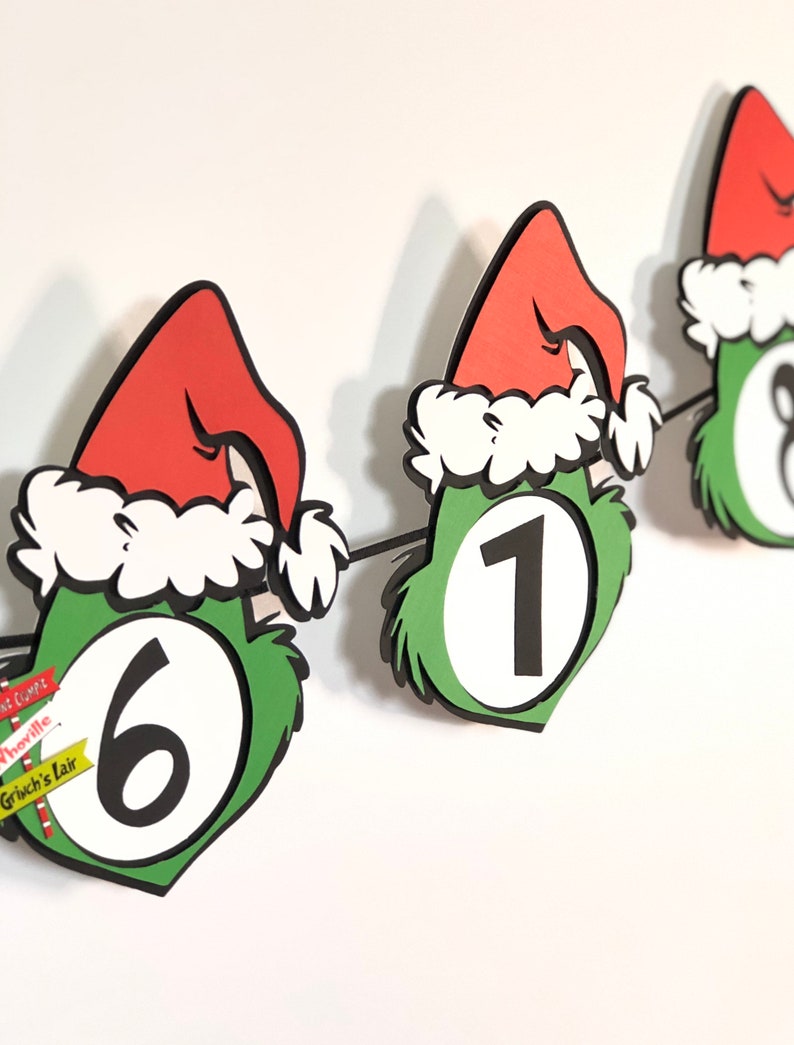 Pode incluir: Uma faixa festiva com recortes de rostos verdes com tema Grinch, com chap&eacute;us de Papai Noel vermelhos e acabamentos brancos. Cada recorte exibe um grande n&uacute;mero preto. A faixa inclui pequenos sinais com texto como "Whoville".