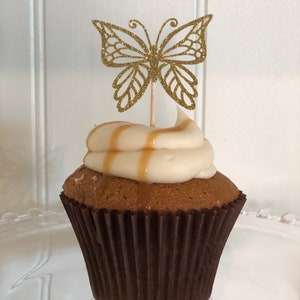 Pu&ograve; includere: Un cupcake con glassa bianca e una spruzzata di caramello, sormontato da un pick a farfalla dorata glitterata. Il cupcake &egrave; in un pirottino di carta marrone su un piatto di vetro trasparente.
