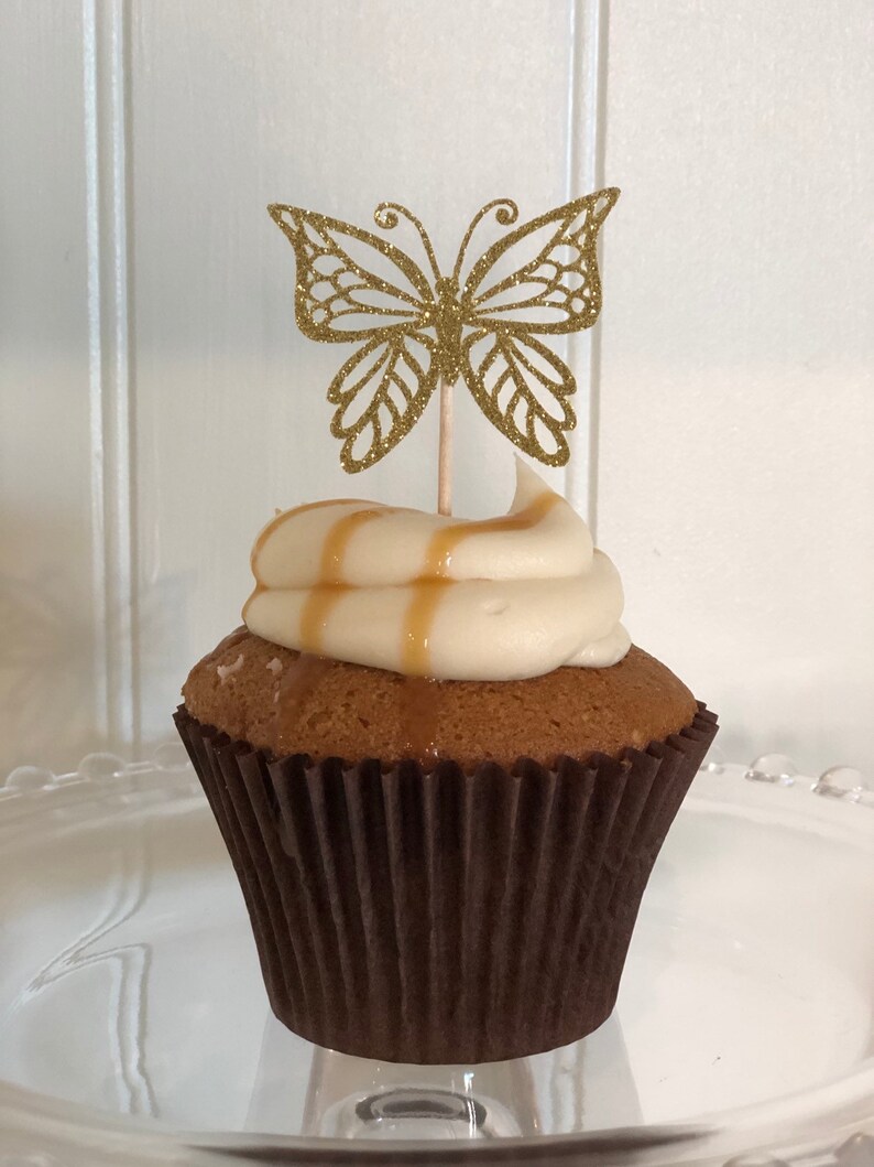 Pu&ograve; includere: Un cupcake con glassa bianca e una spruzzata di caramello, sormontato da un pick a farfalla dorata glitterata. Il cupcake &egrave; in un pirottino di carta marrone su un piatto di vetro trasparente.