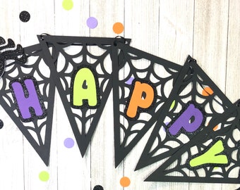 May include: A black Halloween banner with the word "HAPPY" in colorful letters. The banner is decorated with spiderweb cutouts and glittery spiders wearing purple hats. Confetti in various colors is scattered around.