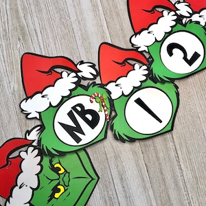 Pode incluir: Uma faixa de Natal festiva com recortes tem&aacute;ticos do Grinch, verdes com chap&eacute;us de Papai Noel vermelhos e acabamentos brancos. A faixa inclui os n&uacute;meros 1 e 2, e as letras NB, juntamente com uma bengala doce. O rosto do Grinch tamb&eacute;m &eacute; vis&iacute;vel.