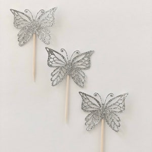 Pu&ograve; includere: Tre decorazioni a forma di farfalla argentata glitterata con bastoncini di legno. Le decorazioni a farfalla presentano intricati disegni traforati e sono disposte diagonalmente su uno sfondo bianco. Ideali per decorare torte o progetti creativi.