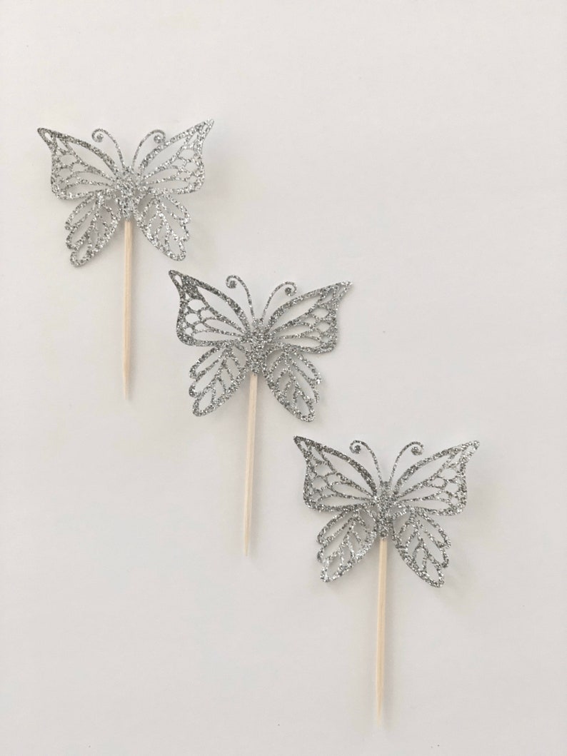 Pu&ograve; includere: Tre decorazioni a forma di farfalla argentata glitterata con bastoncini di legno. Le decorazioni a farfalla presentano intricati disegni traforati e sono disposte diagonalmente su uno sfondo bianco. Ideali per decorare torte o progetti creativi.