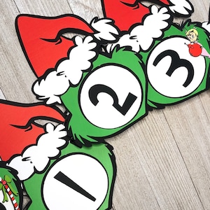 Pode incluir: Uma faixa festiva com c&iacute;rculos verdes com bordas peludas e os n&uacute;meros brancos 1, 2 e 3. Cada c&iacute;rculo &eacute; encimado por um gorro de Papai Noel vermelho com acabamento em pele branca. Uma decora&ccedil;&atilde;o de f&eacute;rias caprichosa.