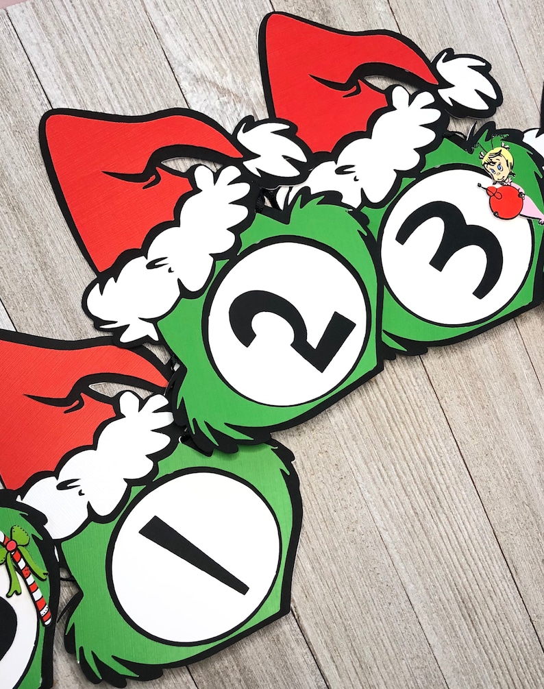 Pode incluir: Uma faixa festiva com c&iacute;rculos verdes com bordas peludas e os n&uacute;meros brancos 1, 2 e 3. Cada c&iacute;rculo &eacute; encimado por um gorro de Papai Noel vermelho com acabamento em pele branca. Uma decora&ccedil;&atilde;o de f&eacute;rias caprichosa.