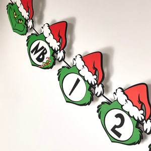 Pode incluir: Uma faixa de Natal extravagante com recortes tem&aacute;ticos do Grinch. A faixa inclui rostos verdes do Grinch, chap&eacute;us de Papai Noel vermelhos com acabamentos brancos e recortes numerados e com letras. A faixa &eacute; amarrada com uma corda preta.