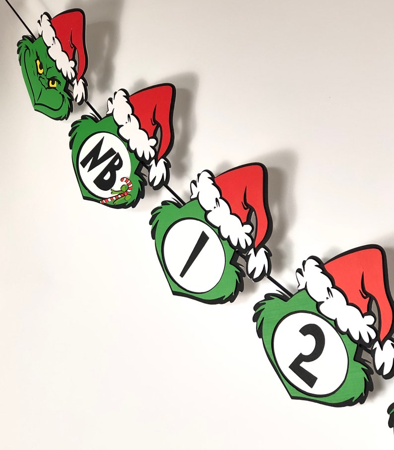 Pode incluir: Uma faixa de Natal extravagante com recortes tem&aacute;ticos do Grinch. A faixa inclui rostos verdes do Grinch, chap&eacute;us de Papai Noel vermelhos com acabamentos brancos e recortes numerados e com letras. A faixa &eacute; amarrada com uma corda preta.