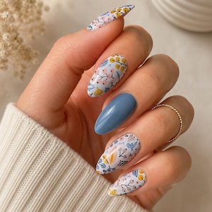 May include: Almond-shaped nails featuring a light blue base with a delicate botanical pattern in shades of yellow, pink, and dark blue, accented by a single solid blue nail and a thin gold ring on the ring finger, worn with a white ribbed jumper.