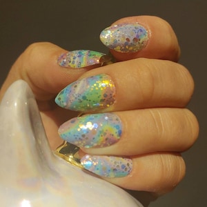 May include: Oval-shaped, iridescent glitter nail art in pastel colors. The nails feature a base of light blue, pink, and yellow with scattered glitter. The hand is holding a white object with a gold accent.