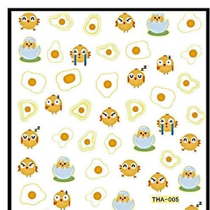 Puede incluir: Una hoja de pegatinas para uñas con pollitos amarillos y huevos. Las pegatinas están sobre un fondo blanco y tienen una variedad de diseños, incluyendo pollitos saliendo de los huevos, pollitos con diferentes expresiones y huevos con yema dentro. El texto "THA-005" está impreso en la hoja.