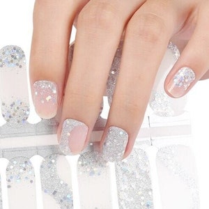 Può includere: Un foglio di strisce di smalto per unghie glitter argento con un design a spirale bianco e argento. Le strisce sono disposte in fila e sono pronte per essere applicate sulle unghie.