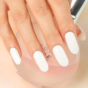 Peut inclure: Une main avec de longs ongles blancs de forme ovale. Une bague en argent avec un motif de papillon est à l'annulaire. La main tient un flacon de parfum transparent avec un bouchon argenté.