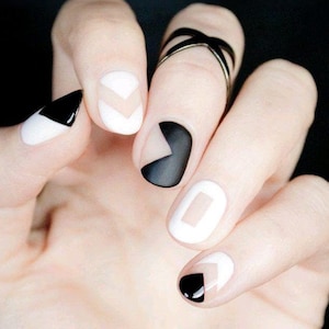 May include: A hand displaying a contemporary manicure with black, white, and nude geometric patterns. The nails are decorated with triangles and chevrons. A black ring with a crisscross design is on a finger.