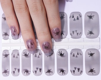 May include: Gray nail wraps with a spooky Halloween theme. Each wrap features a ghostly hand print design in black. The nail wraps are shown on a hand with painted nails, showcasing the design.